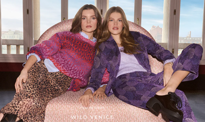 2 Frauen sitzen in einem hellrosa Sessel und schauen in die Kamera. Eine Frau trägt einen Rock mit Leopardenprint, ein hellblaues Hemd und einen rosa Pullover. Die andere Frau trägt einen violetten Anzug mit Muster und schwarze Schuhe